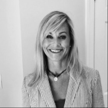 Smiling woman with long blonde hair wearing a light pinstripe blazer and a necklace, posed indoors against a light background.