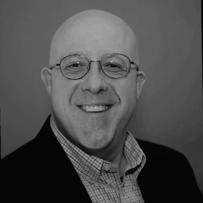 Smiling man with glasses and a bald head wearing a dark blazer over a checkered shirt, posed against a plain background.
