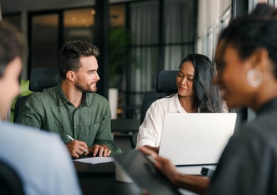 Four colleagues in a modern office meeting, with two people in focus discussing notes and using a laptop, while others listen in the foreground.
