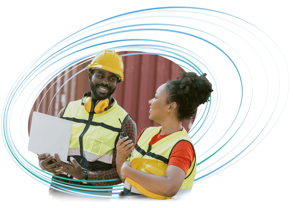 Two logistics workers in safety vests and hard hats conversing near shipping containers; one holds a laptop and the other a walkie-talkie.