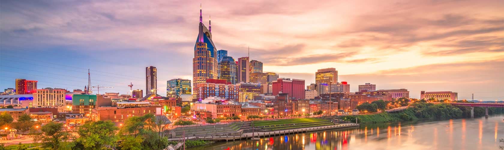 Panoramic view of downtown Nashville at sunset, featuring the iconic AT&T building, illuminated city lights, and reflections on the Cumberland River.